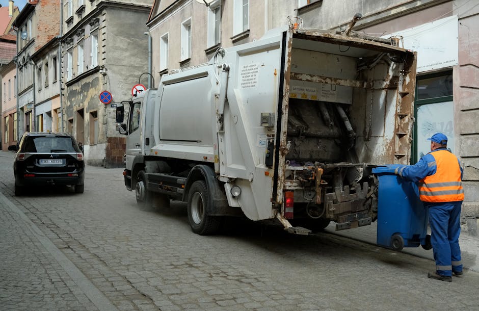 A large collection of mixed waste and rubbish overflowing from multiple black and gray recycling bins, with discarded packaging, cardboard boxes, plastic bags, and paper scattered on the pavement in f