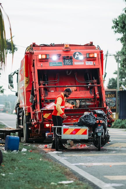 A sanitation worker wearing a high-visibility red and yellow uniform operates a red refuse collection truck during a rubbish collection task on a roadside. The worker is standing behind a waist-high m